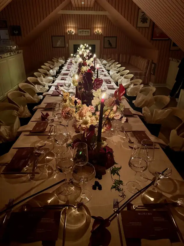 Long dinner table set beneath a pitched ceiling with candlelight and deep red florals
