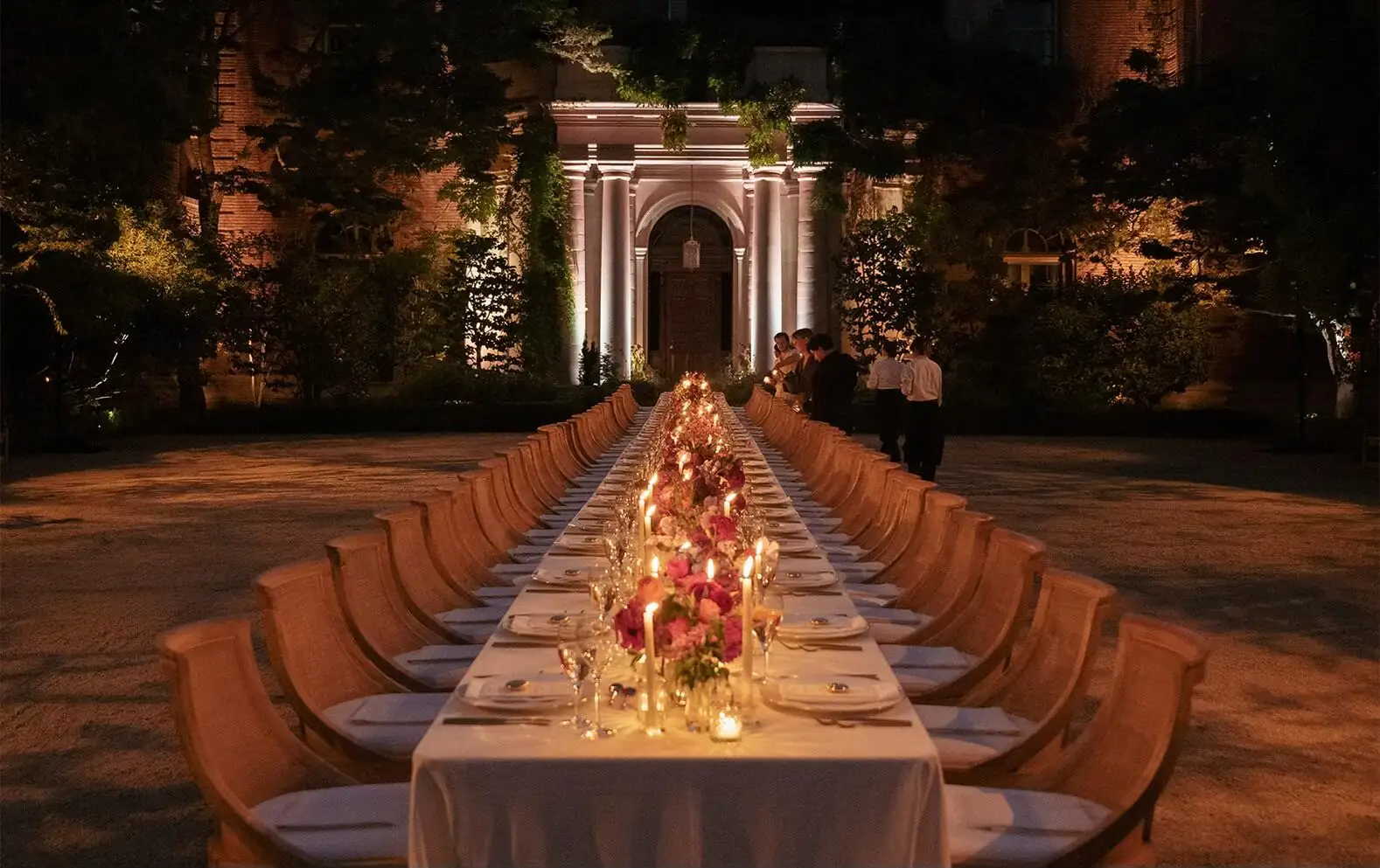 A long candlelit dinner table set beneath an illuminated stone archway at night