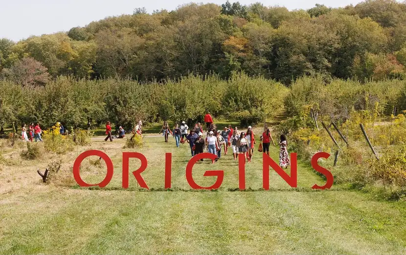 Guests gathered around oversized ORIGINS letters set in an autumn orchard