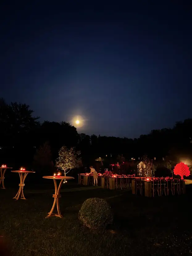 The orchard dinner at nightfall, lit in red beneath a rising moon